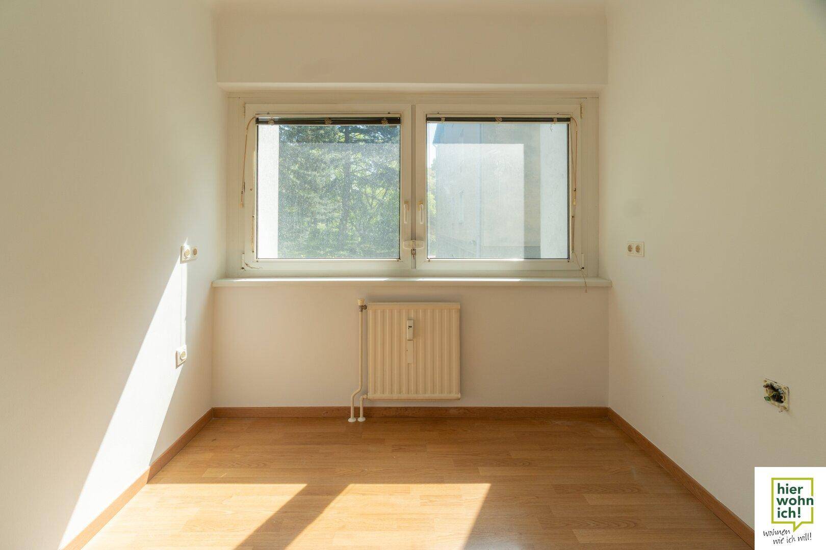 Freundliches Zimmer mit großem Fenster, Parkettboden und einem Heizkörper.