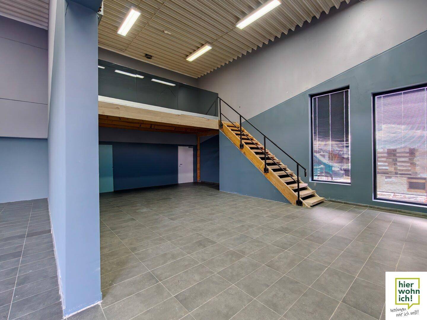 Großer Gewerberaum mit hohen Decken, Fliesenboden, einer Holztreppe und einer Galerieebene.