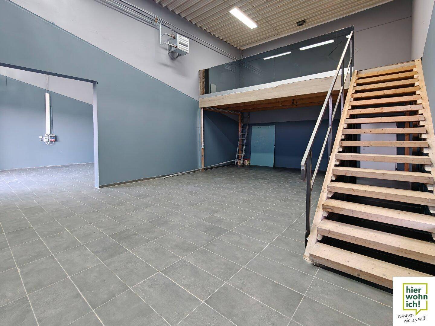 Geräumiger Gewerberaum mit Fliesenboden, hoher Decke und einer Holztreppe, die zur Galerie führt.