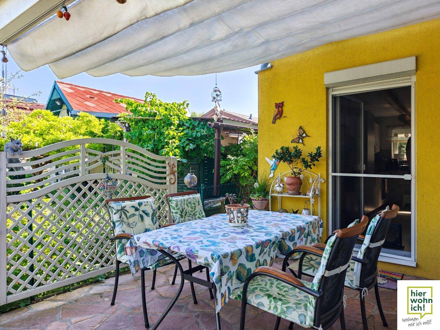 Überdachte Terrasse mit Esstisch und Stühlen, umgeben von einem dekorativen Zaun und Pflanzen.