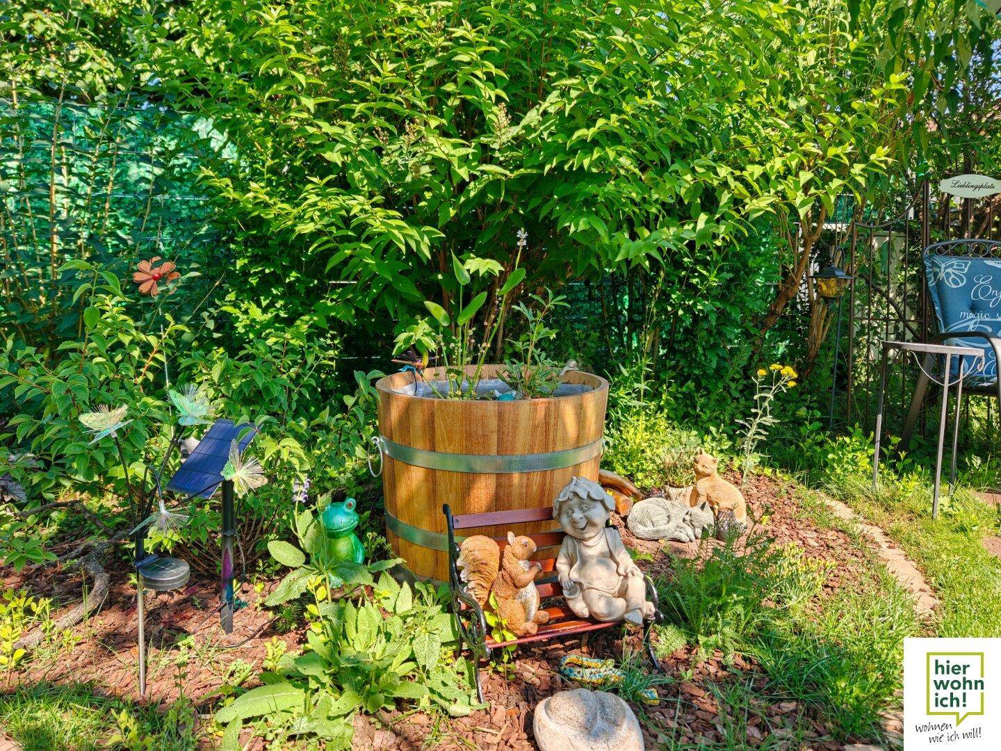 Detailansicht eines Gartenteils mit Holzfass, Zwergenfiguren und üppiger Bepflanzung.