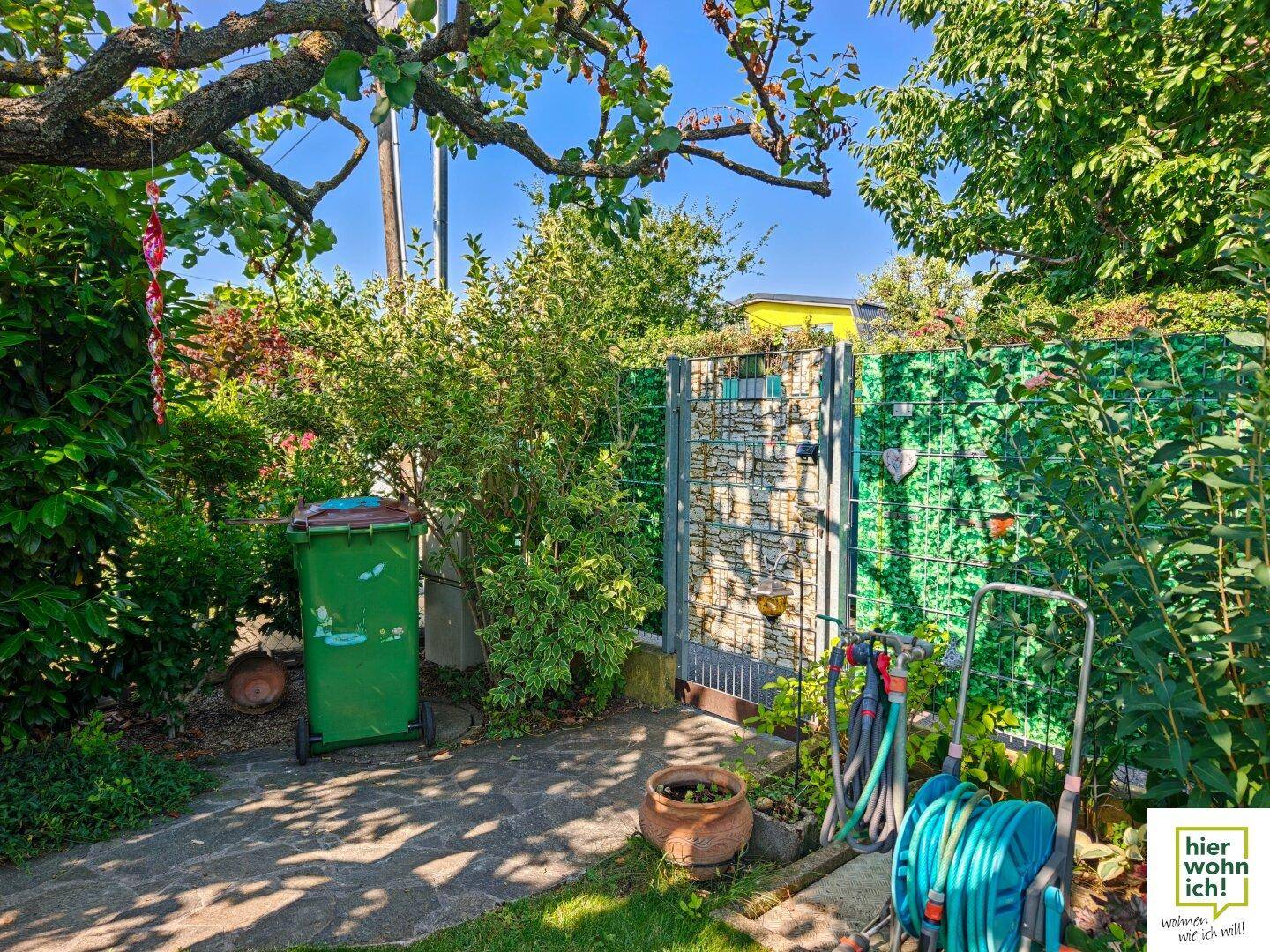 Gartenbereich mit grüner Mülltonne, Gartenschlauch und einem dekorativen Gartentor.