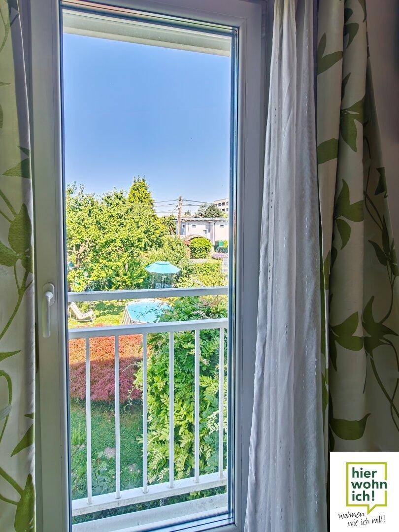 Blick aus dem Fenster auf den grünen Garten und einen kleinen Balkon mit weißem Geländer.