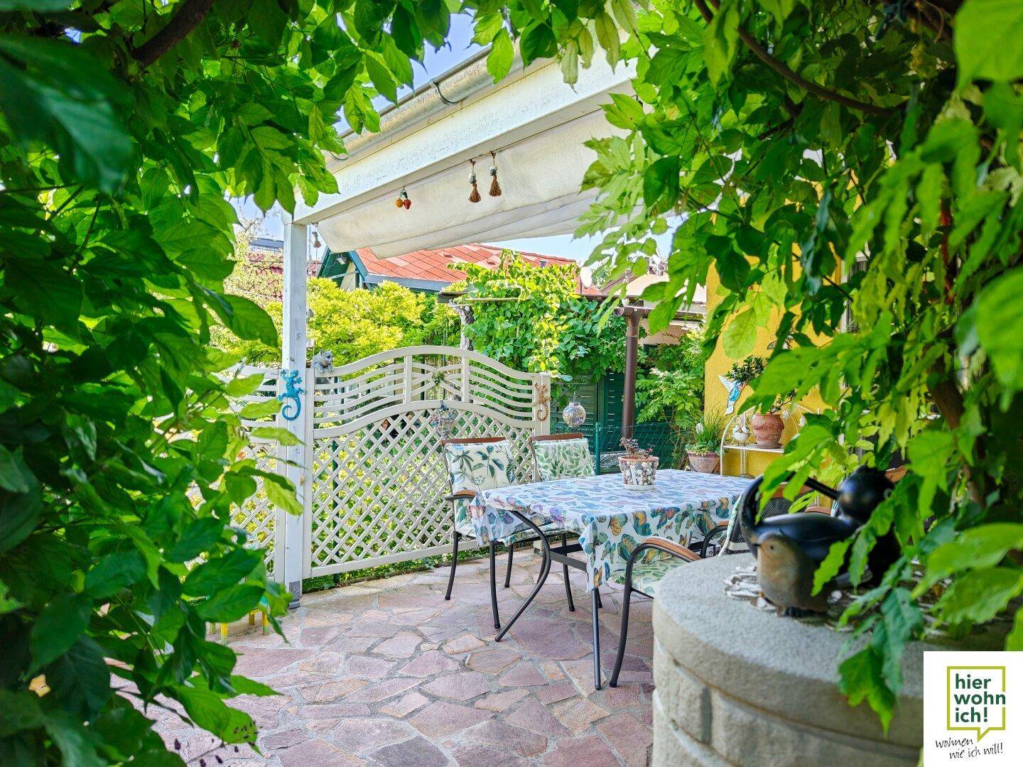 Überdachte Terrasse mit Esstisch und Stühlen, umgeben von üppigem Grün, ideal für Mahlzeiten im Freien.