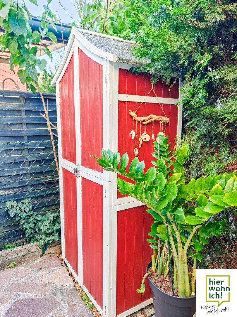 Kleiner rot-weißer Gartenschuppen, umgeben von Pflanzen, bietet Stauraum für Gartengeräte.