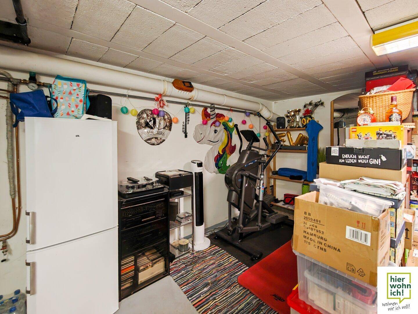Kellerraum mit Kühlschrank, Regalen und Lagerboxen, sowie einem Fitnessgerät.