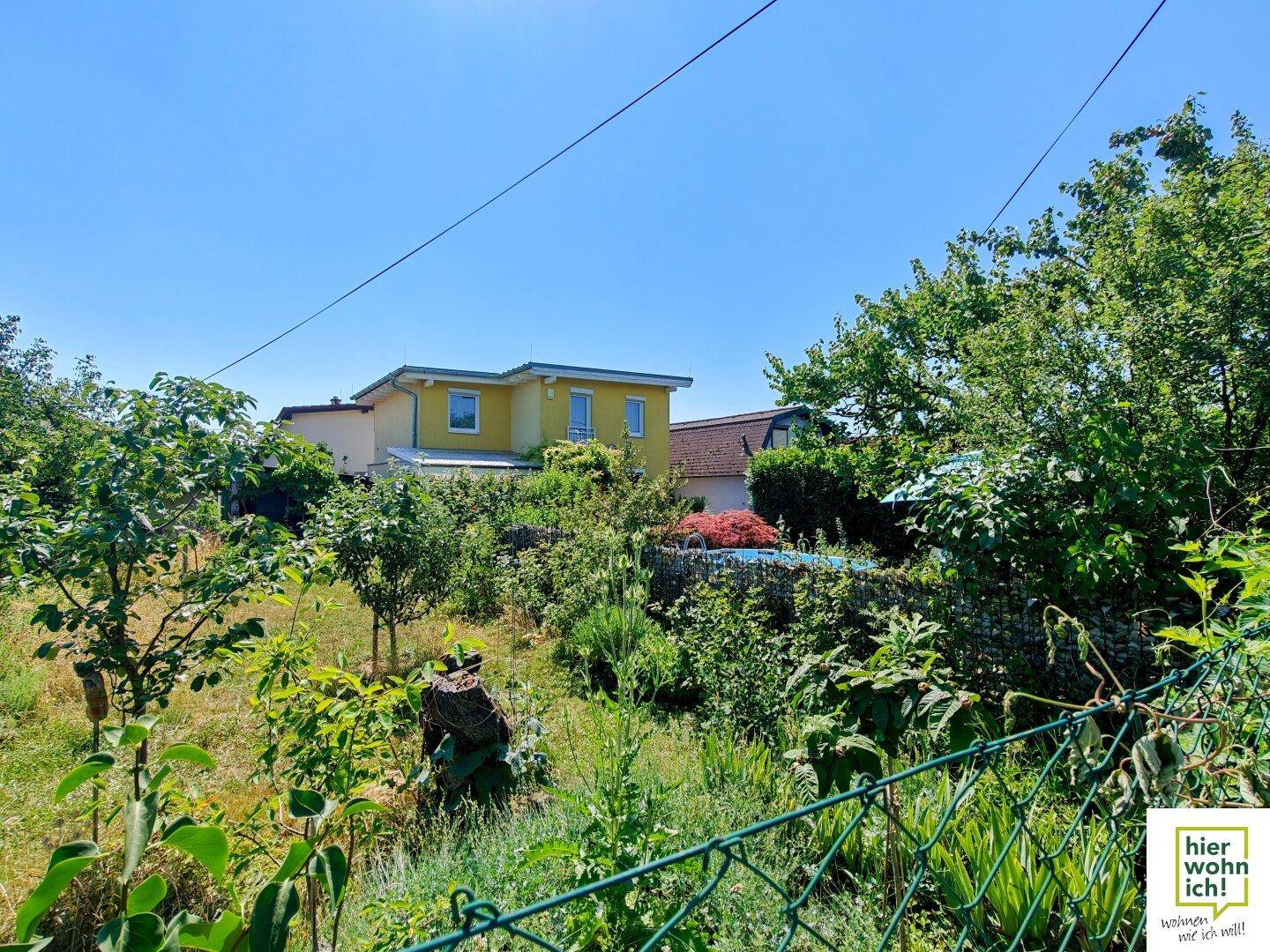 Weitläufiger Blick auf das gelbe Haus, umgeben von üppigem Grün und Bäumen unter blauem Himmel.