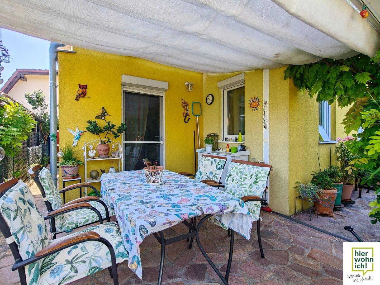 Überdachte Terrasse mit Esstisch, Stühlen und bunten Dekorationen an der gelben Hauswand.