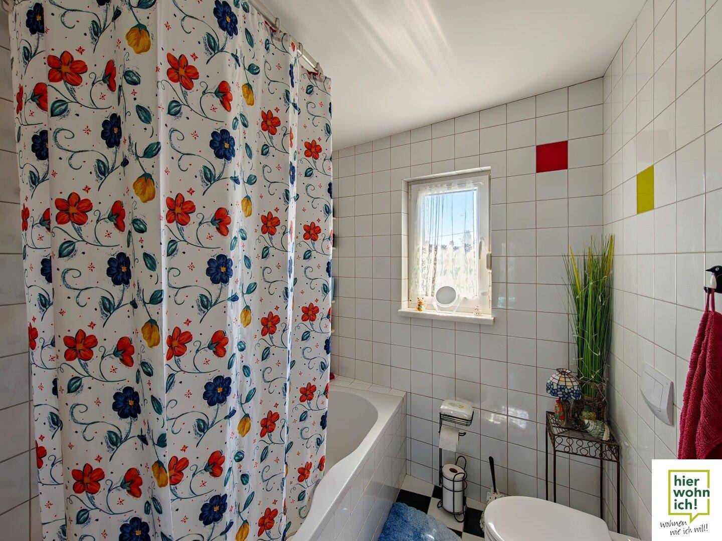 Helles Badezimmer mit Badewanne, farbenfrohem Duschvorhang und weißen Fliesen mit Akzenten.