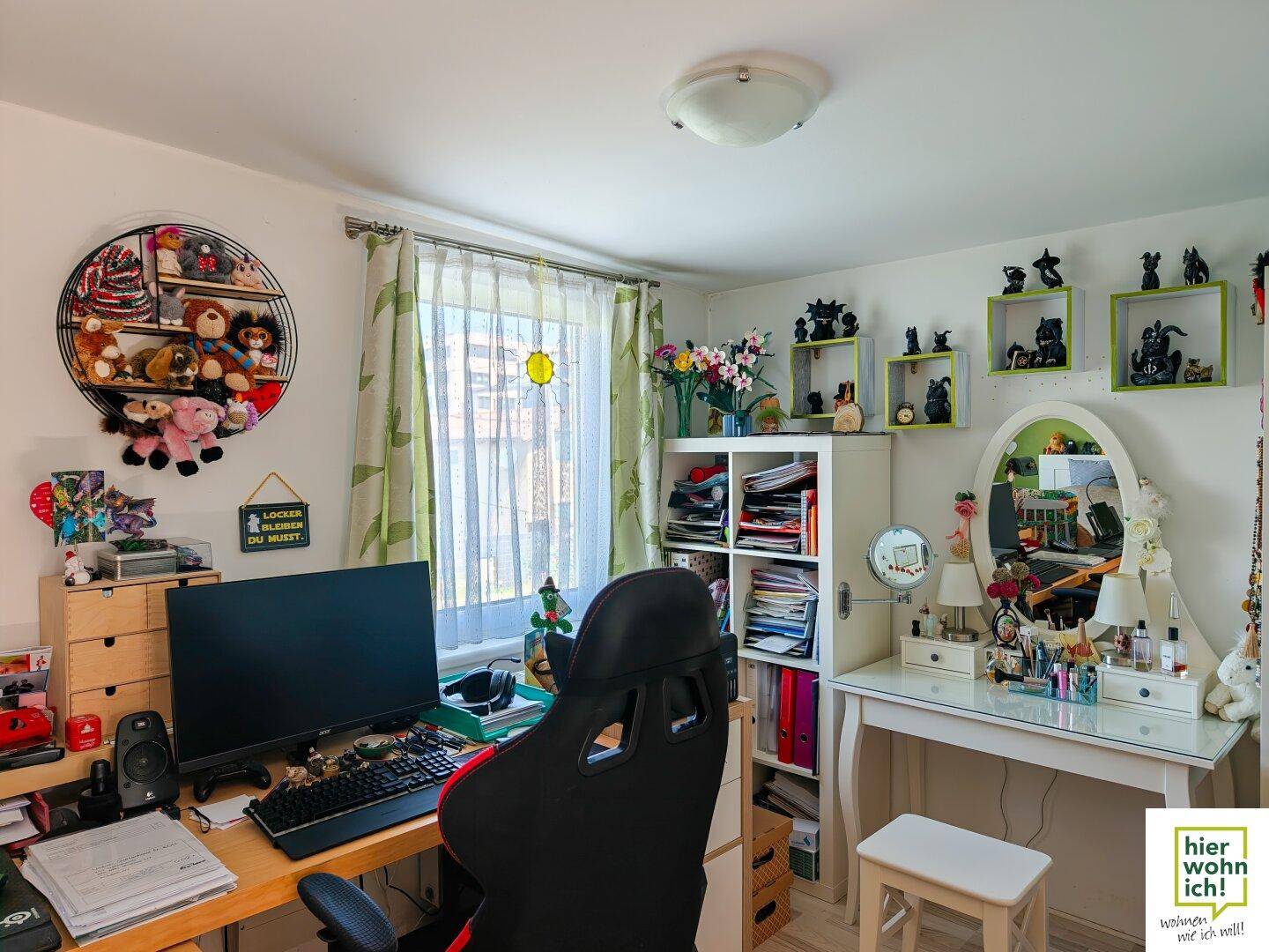 Helles Arbeitszimmer mit Schreibtisch, Computer, Regalen und einem Schminktisch am Fenster.