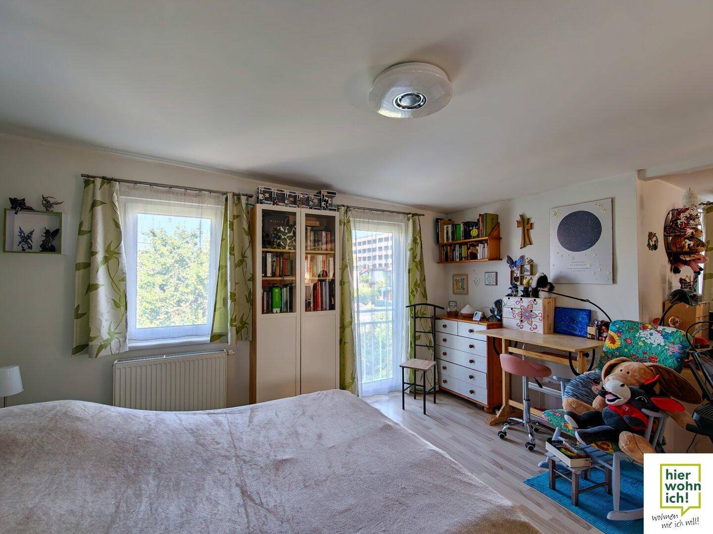 Schlafzimmer mit Bett, Schreibtischbereich, Bücherregalen und Zugang zu einem Balkon.