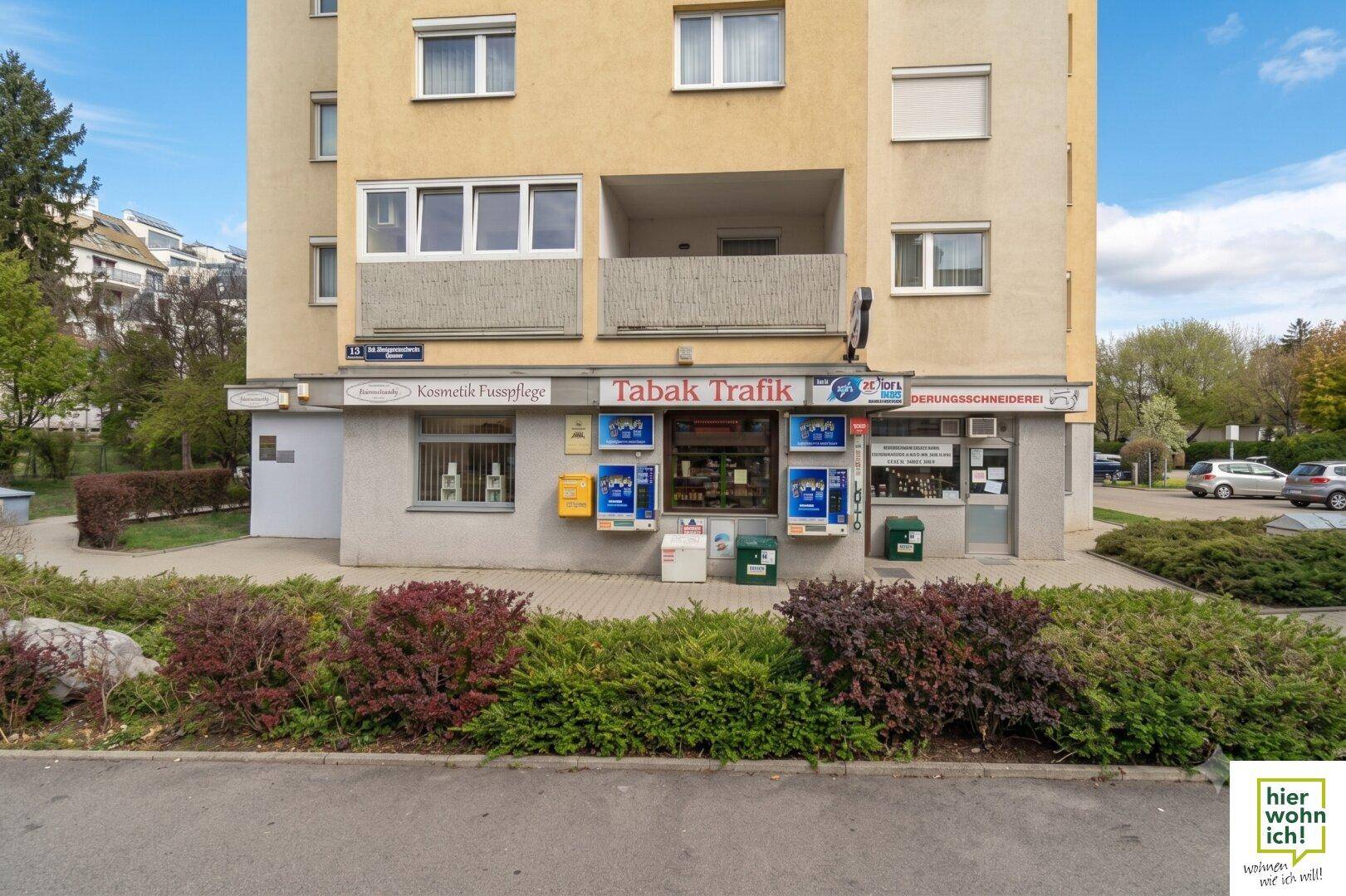 Erdgeschossfassade eines Gebäudes mit kleinen Geschäften und Wohnungen darüber, umgeben von Grünflächen.