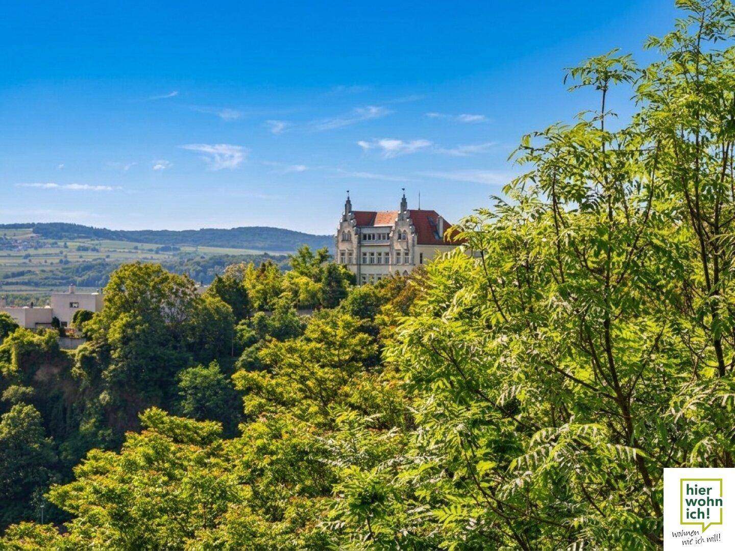 Historisches Gebäude auf einem Hügel, umgeben von dichtem Grün und Bäumen unter blauem Himmel.