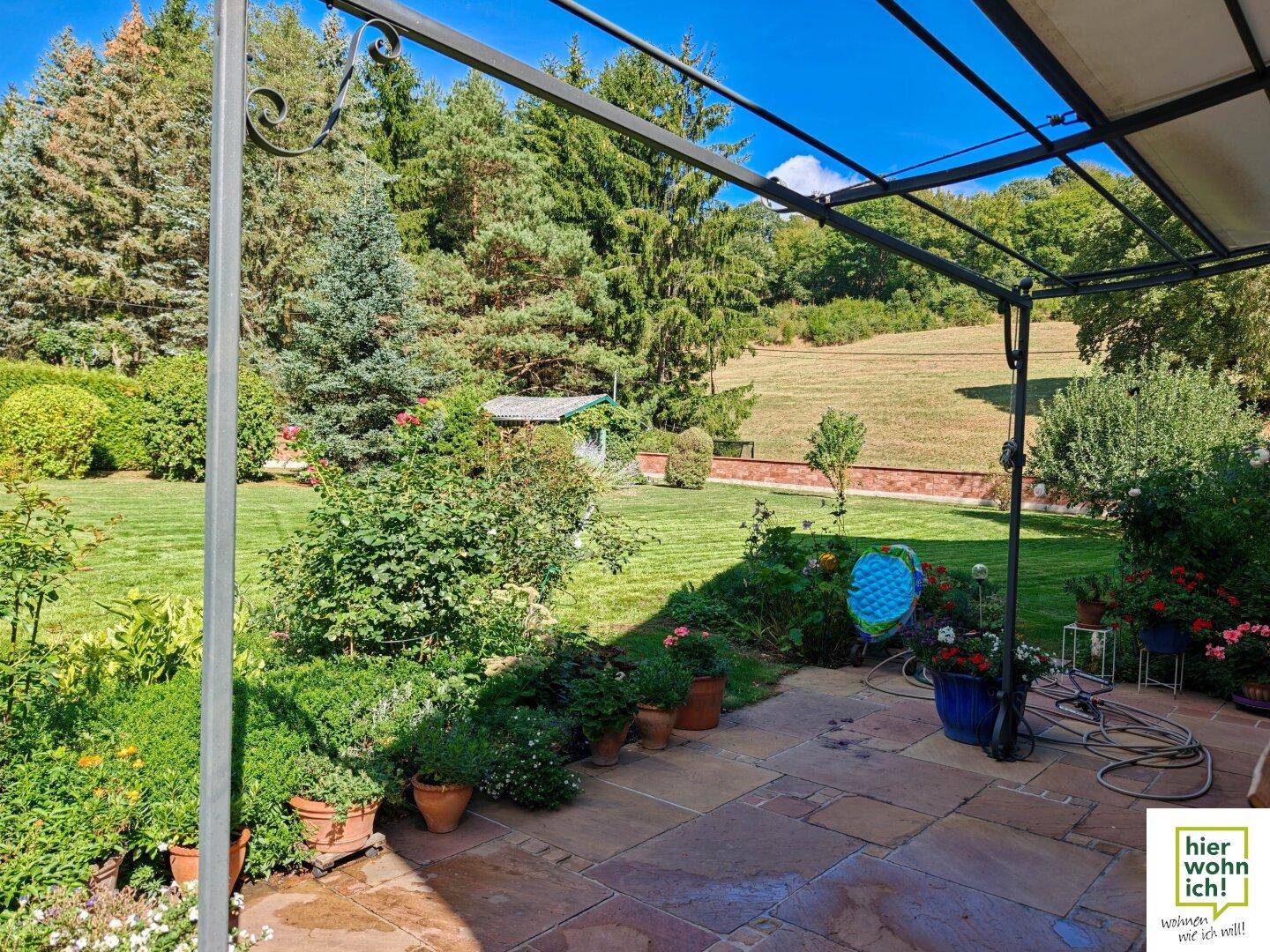 Geräumige Steinterrasse mit Pergola, Blick auf den großen Garten und die umliegende Natur.