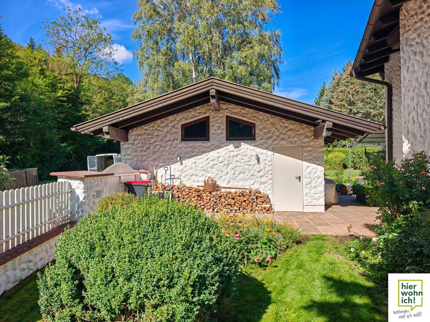 Nebengebäude mit weißer Fassade und Holzlager, umgeben von einem gepflegten Garten.