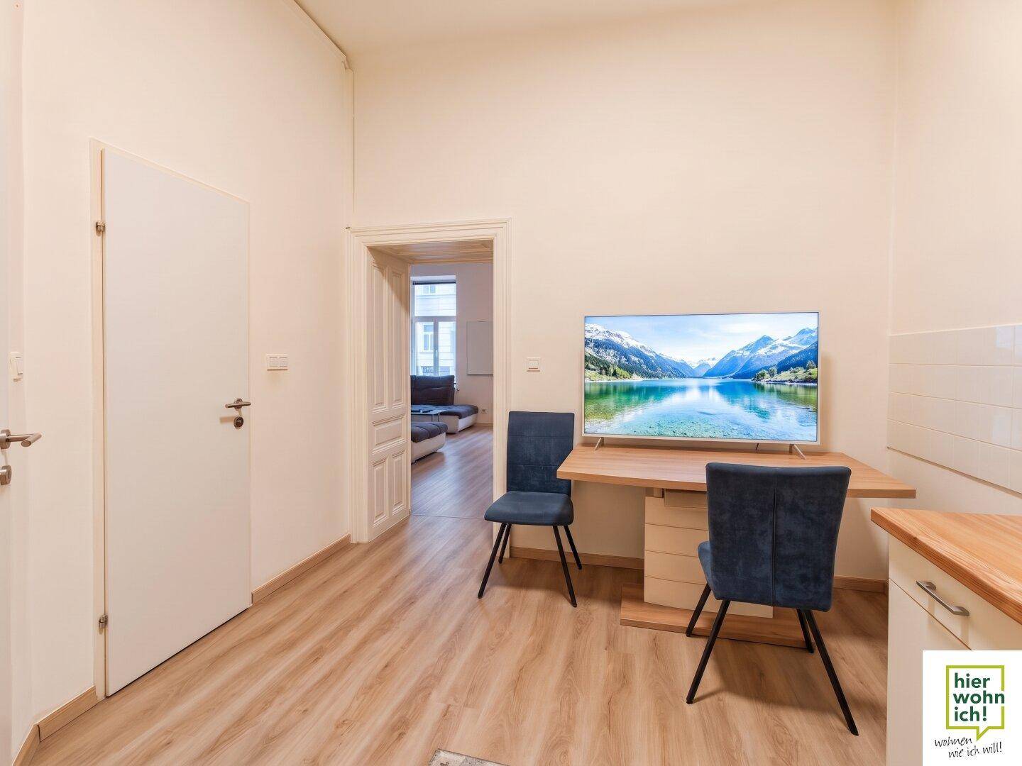 Wohnbereich mit kleinem Esstisch, zwei Stühlen und einem Fernseher, der eine Berglandschaft zeigt.