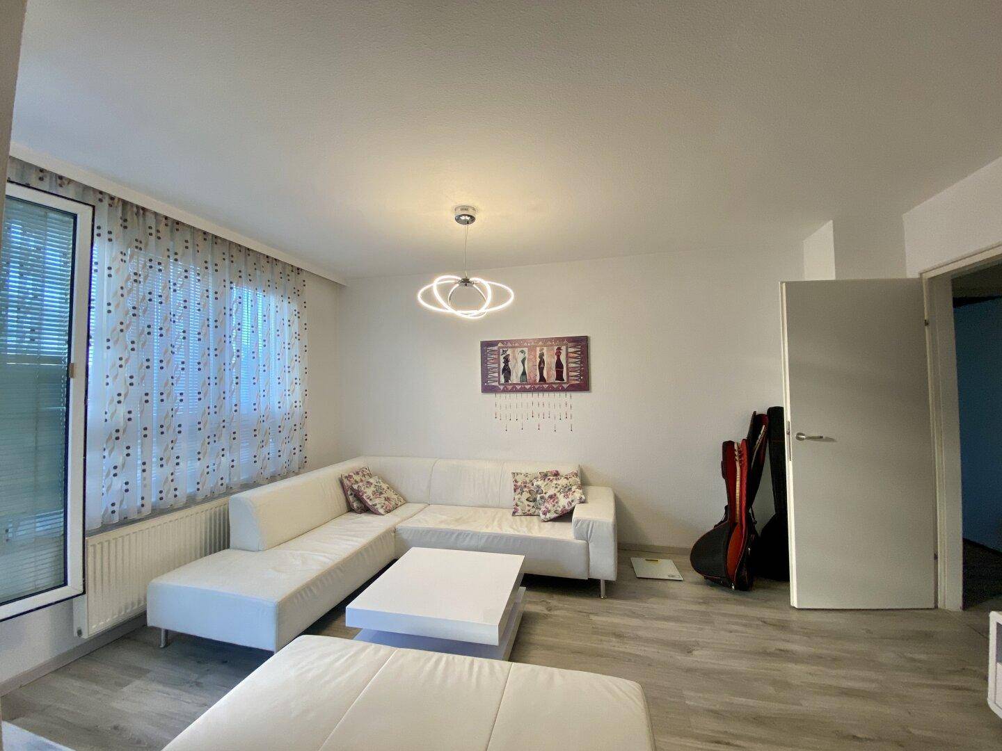 Gemütliches Wohnzimmer mit weißem Ecksofa, modernem Licht und Zugang zum Balkon.