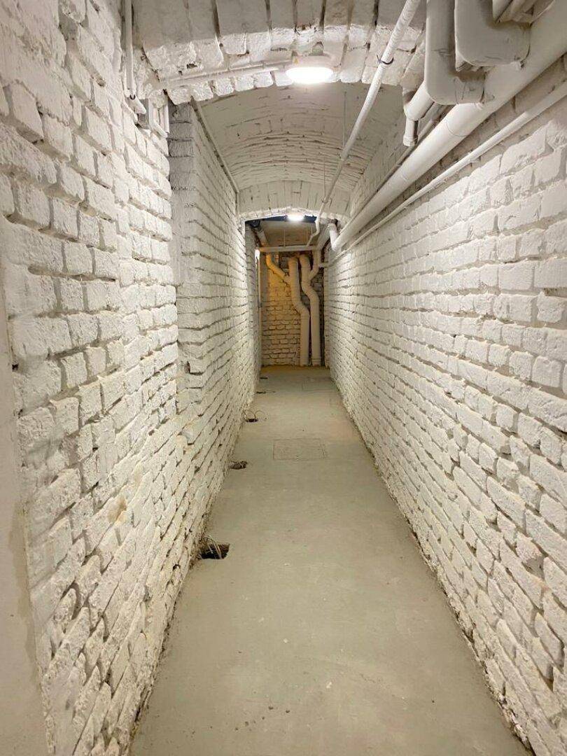 Langer Kellergang mit weiß gestrichenen Ziegelwänden und sichtbaren Rohren an der Decke.