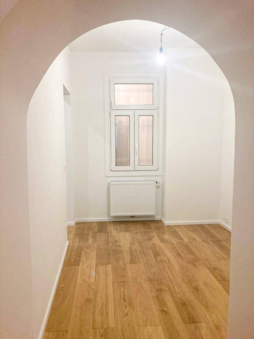 Lichtdurchfluteter Raum mit Holzfußboden und Fenster, zugänglich durch einen eleganten Bogen.