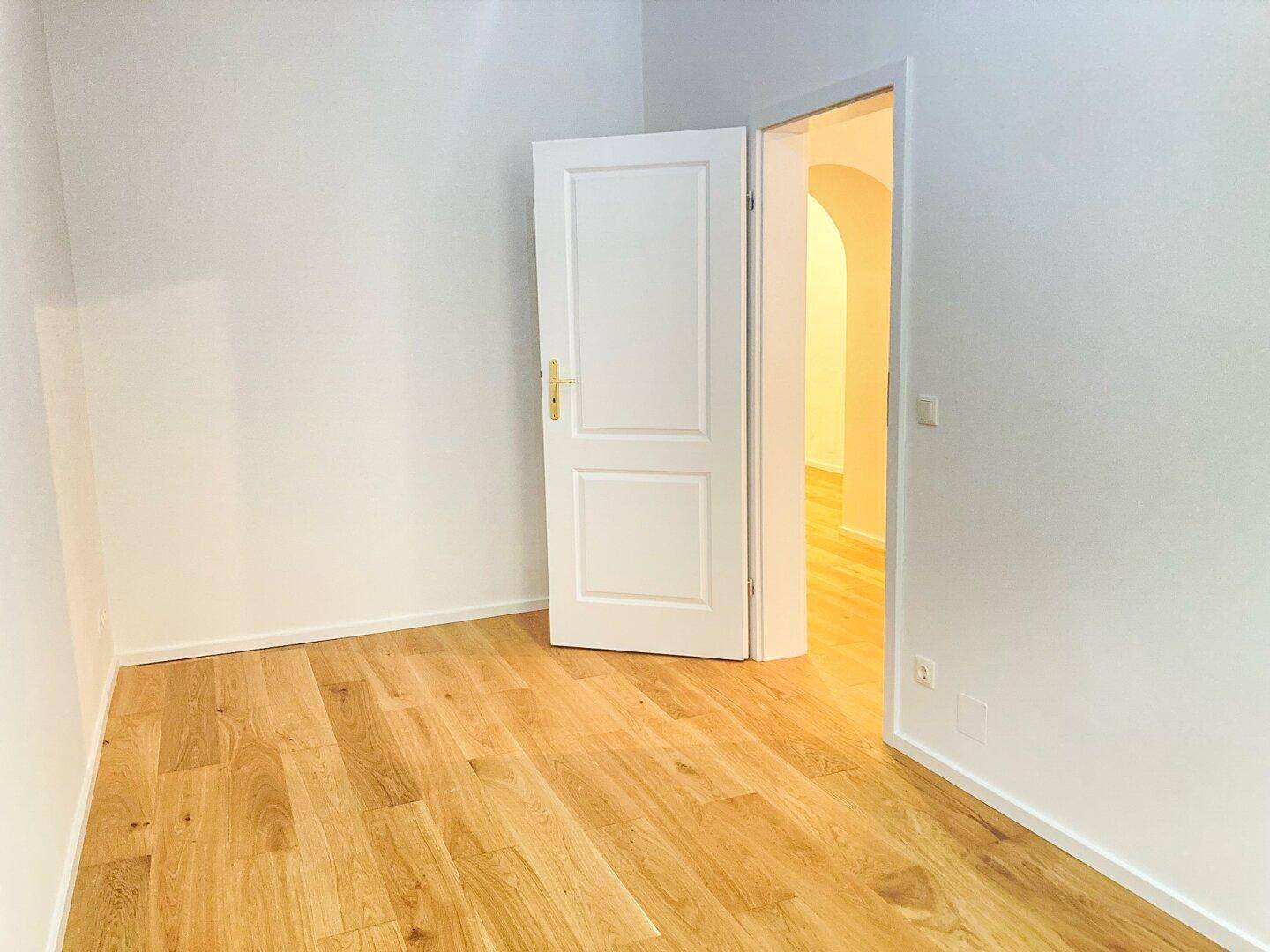Leeres Zimmer mit hellem Holzfußboden und offener Tür, bietet Potenzial für individuelle Einrichtung.