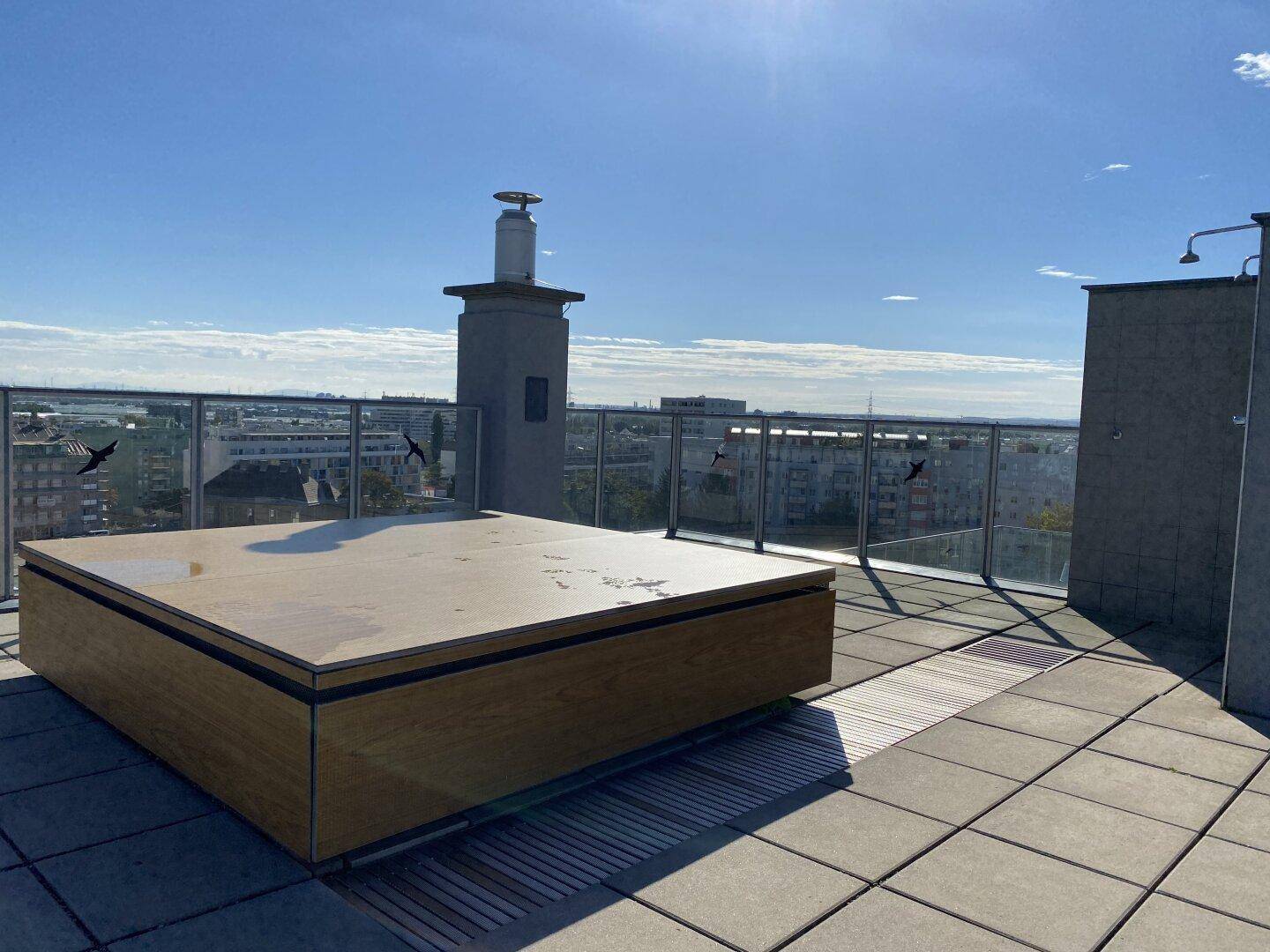 Sonniger Dachbereich mit großem Holzelement, möglicherweise ein Whirlpool, und Stadtpanorama.