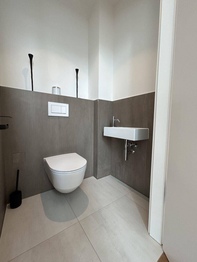 Modernes Gäste-WC mit wandhängender Toilette, kleinem Waschbecken und grauen Fliesen.