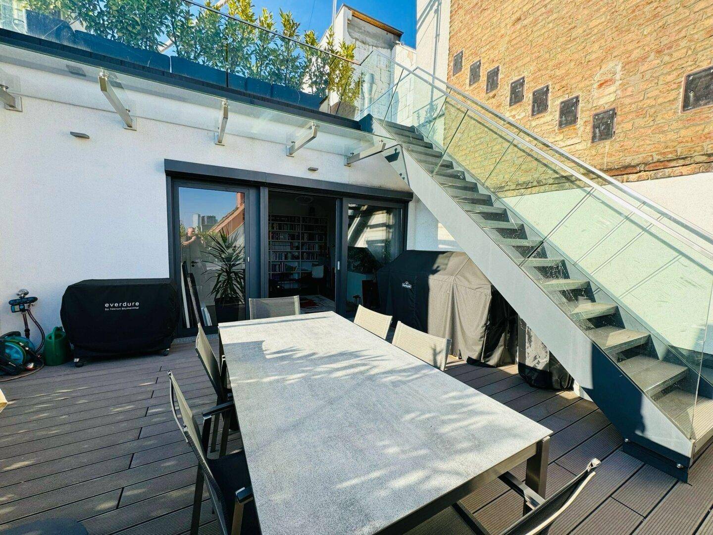 Sonnige untere Dachterrasse mit Esstisch und moderner Treppe zur oberen Ebene.