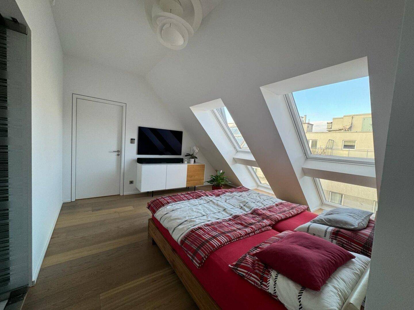 Helles Schlafzimmer unter einer Dachschräge mit Holzboden, Fernseher und großen Fenstern.