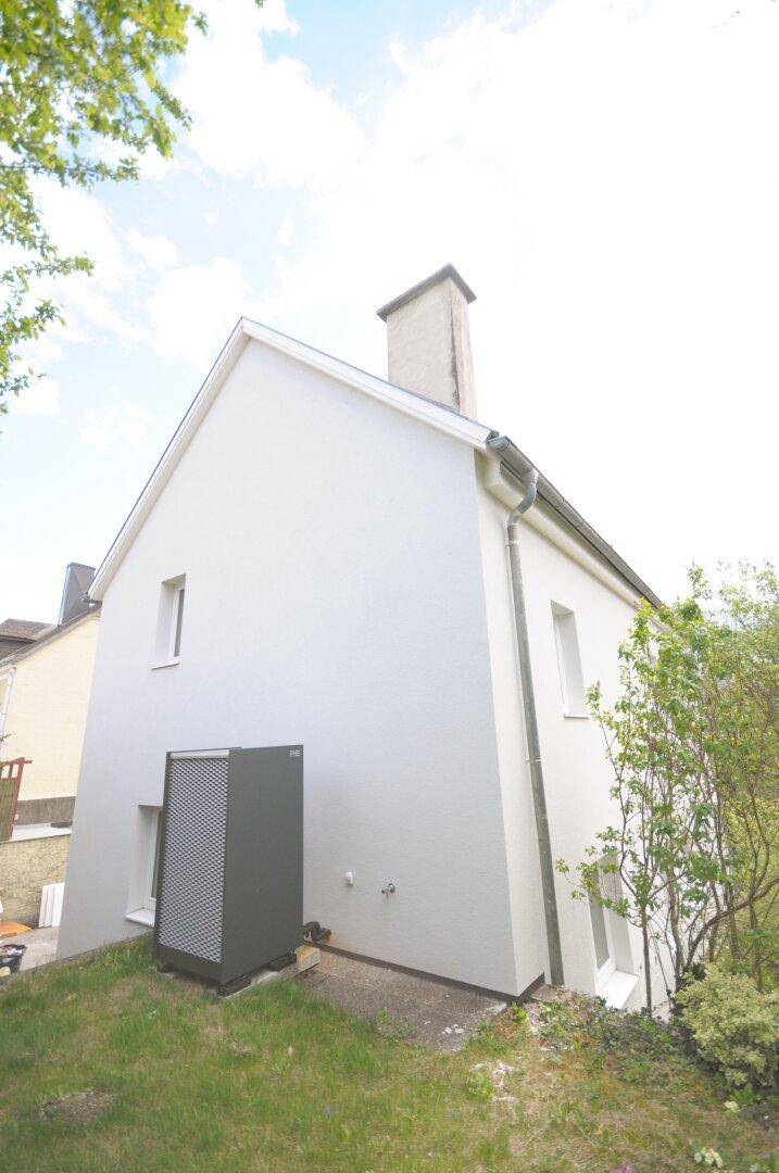 Seitenansicht des weißen Hauses mit Wärmepumpe und grünem Rasen im Gartenbereich.