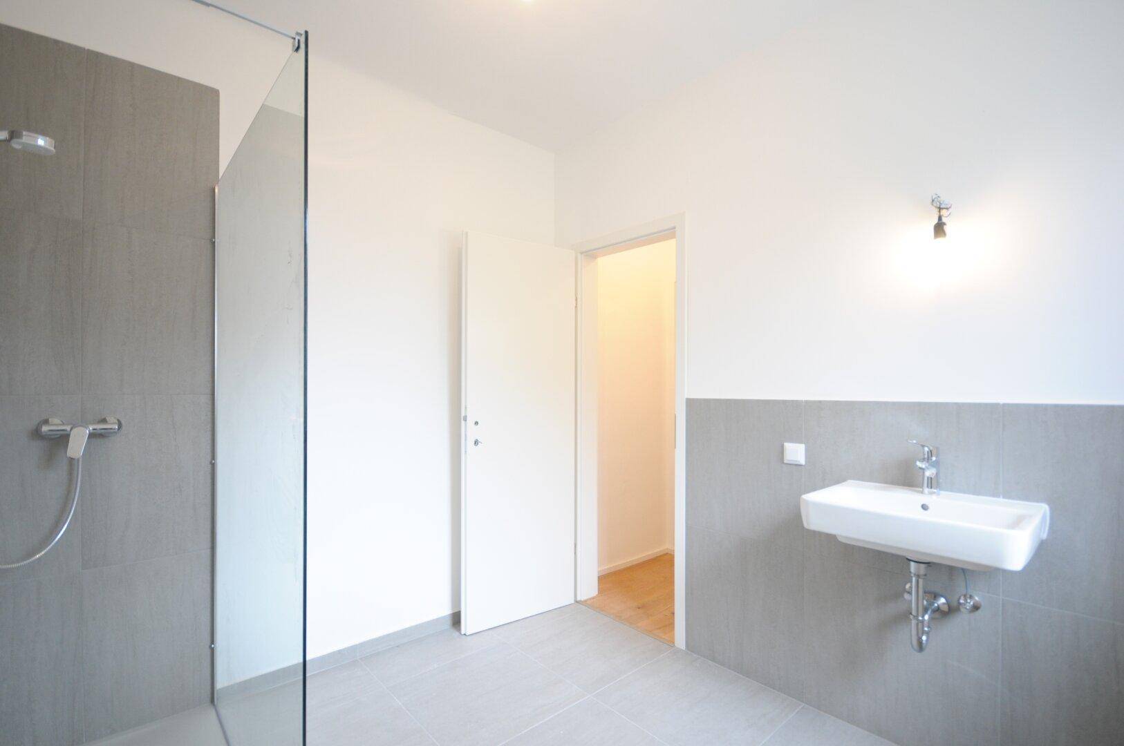 Helles Badezimmer mit moderner Dusche, grauen Fliesen und einem Waschbecken.
