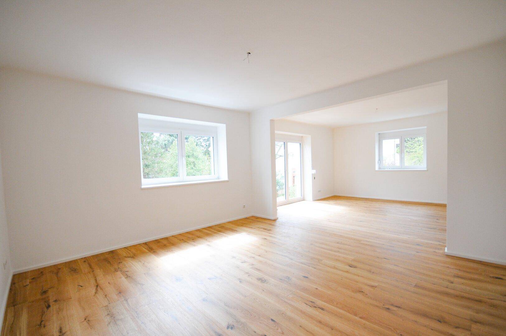 Geräumiges Wohnzimmer mit hellem Holzboden und großen Fenstern, die viel Tageslicht hereinlassen.