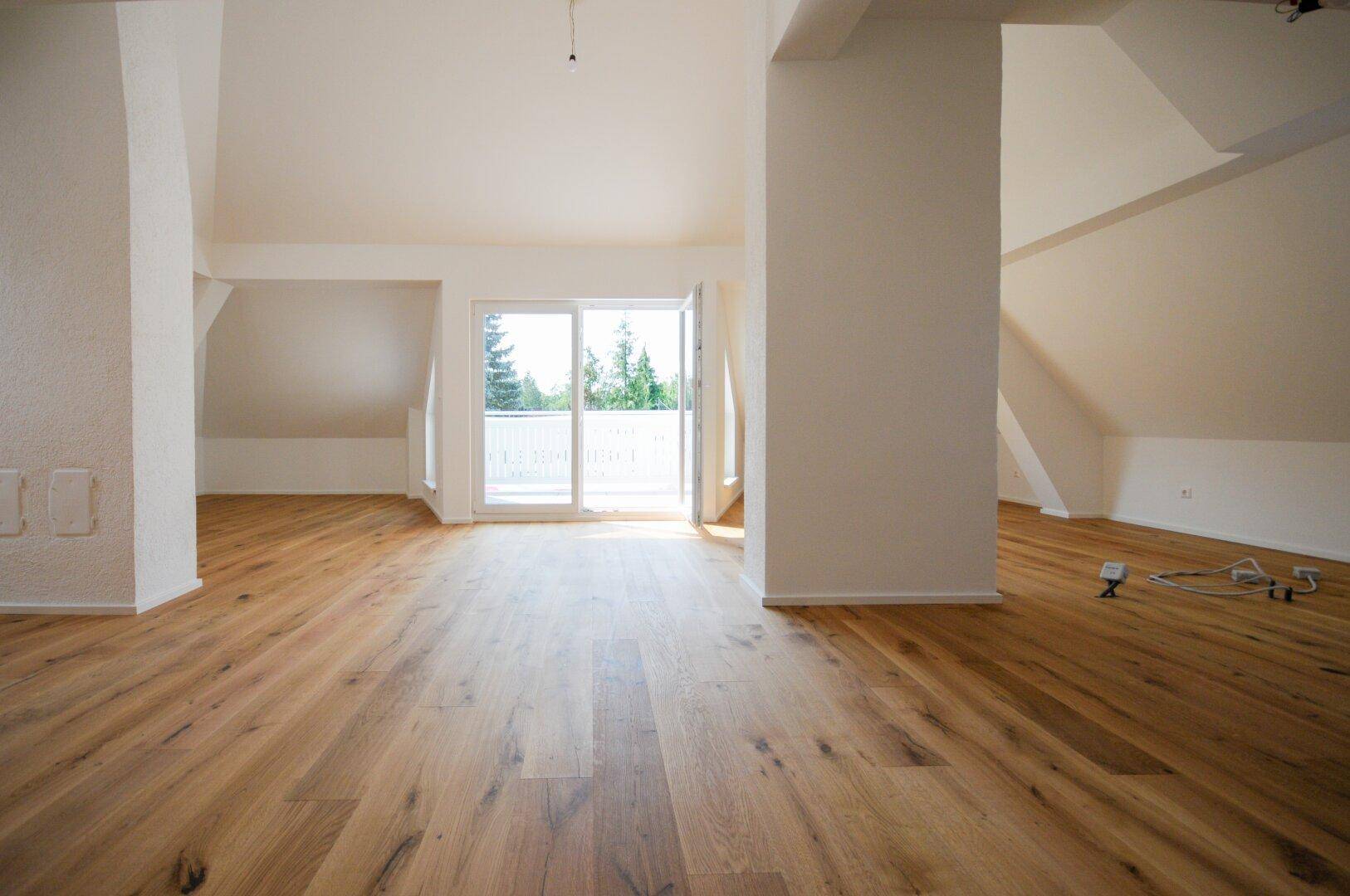 Heller Dachgeschossraum mit Holzboden und Zugang zu einem Balkon, umgeben von grüner Natur.