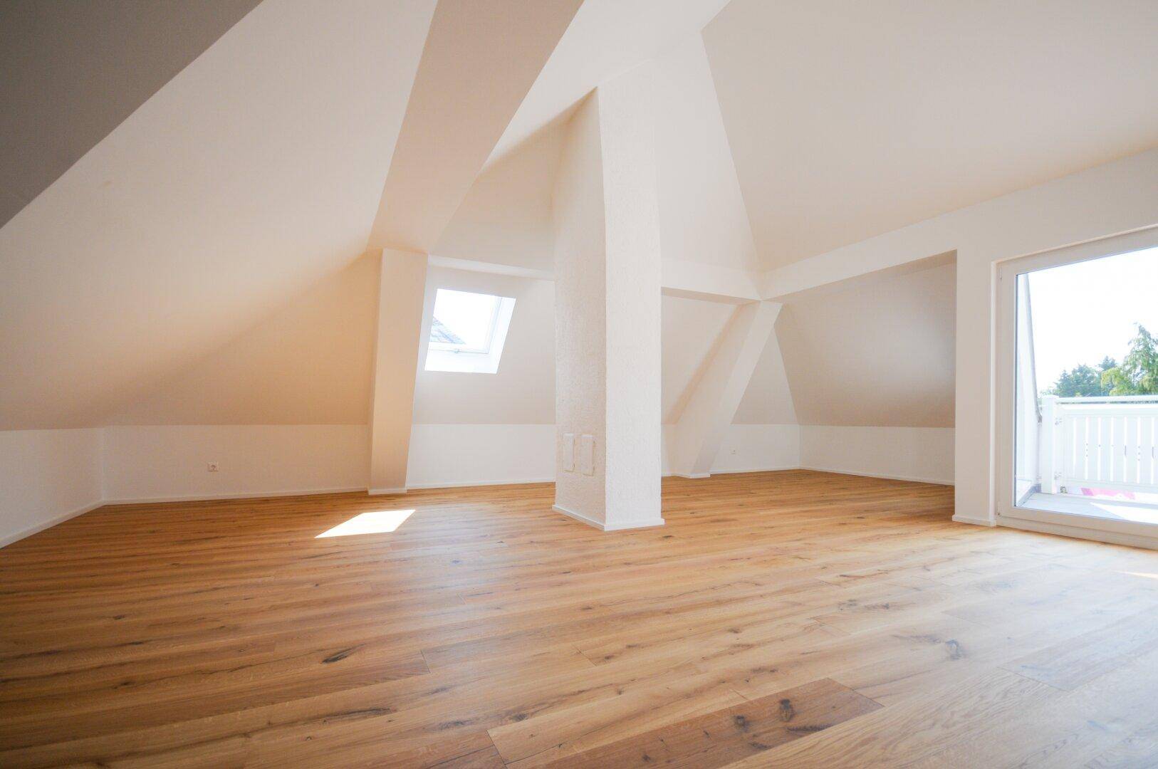 Geräumiger Dachgeschossraum mit Holzboden, Dachfenstern und Zugang zu einem sonnigen Balkon.