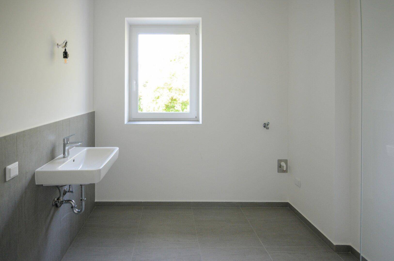 Helles Badezimmer mit Waschbecken, Fenster und Anschluss für Waschmaschine.