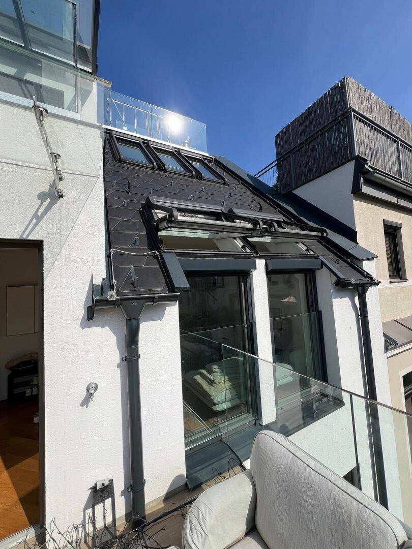 Balkon mit Glasgeländer, Sitzgelegenheiten und Blick auf die Dachlandschaft.