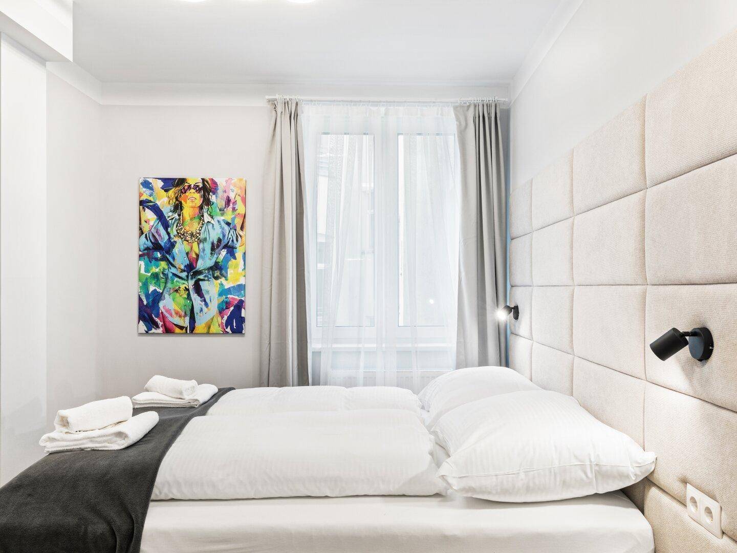 Gemütliches Schlafzimmer mit gepolstertem Kopfteil, Kunstwerk und viel Tageslicht.