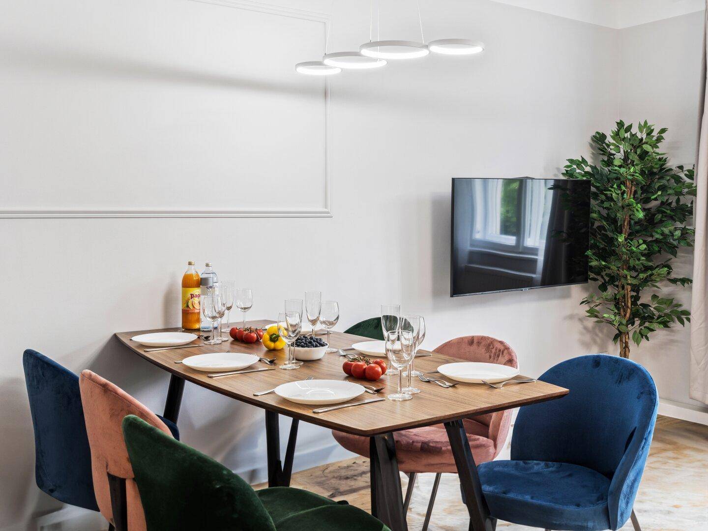 Moderner Essbereich mit farbigen Stühlen, Holztisch und Wandfernseher unter stilvoller Beleuchtung.