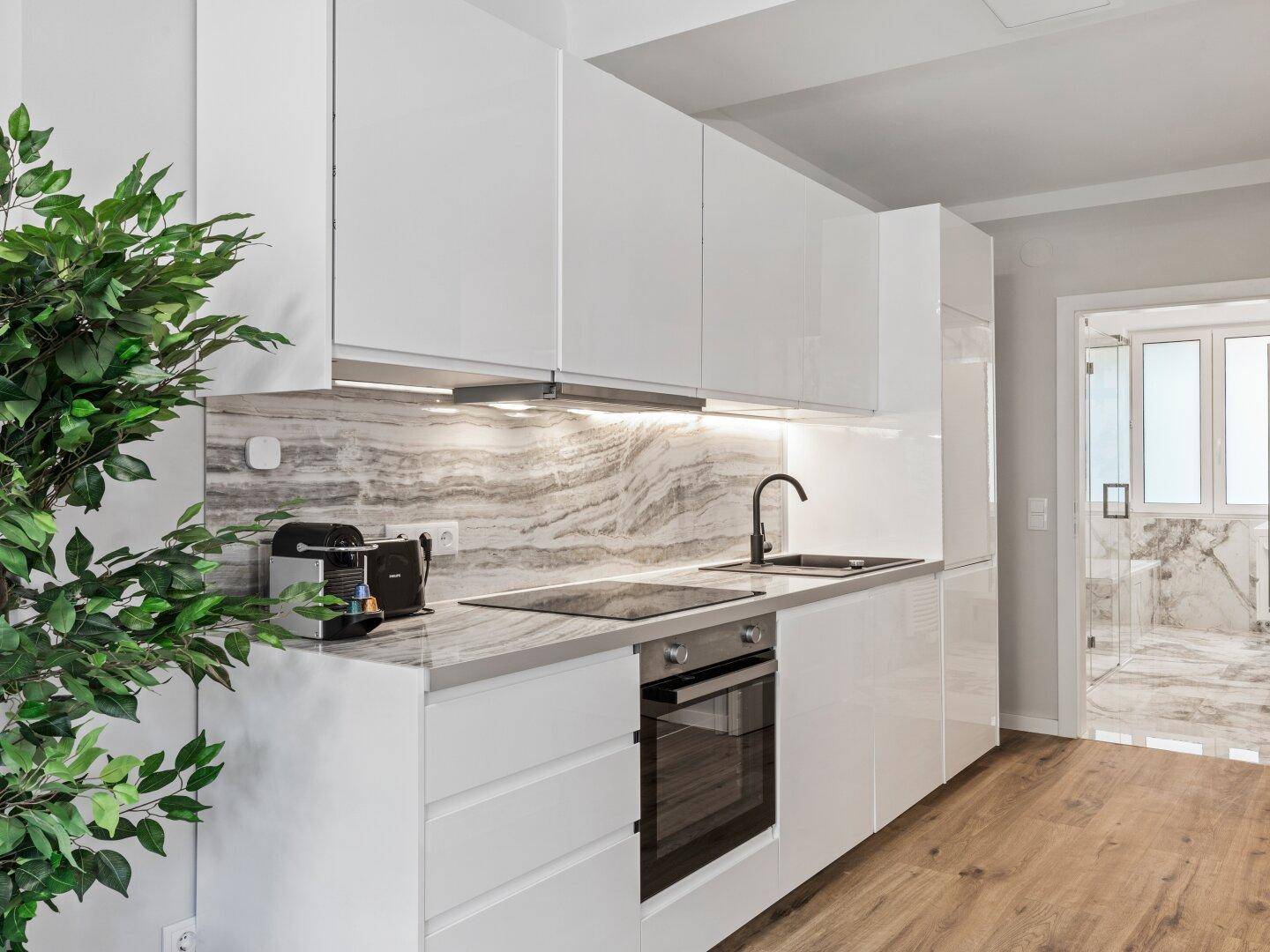 Moderne weiße Küche mit glänzenden Fronten, Marmor-Rückwand und Holzboden, offen zum Flur.