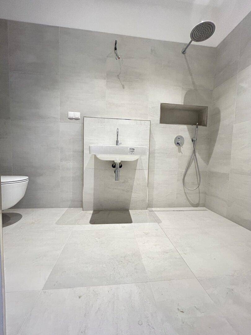 Modernes Badezimmer mit hellgrauen Fliesen, wandhängendem WC und einer geräumigen Walk-in-Dusche.