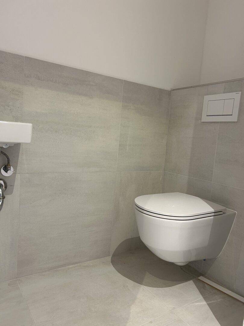Gäste-WC mit wandhängender Toilette und kleinem Waschbecken, komplett gefliest in hellem Grau.
