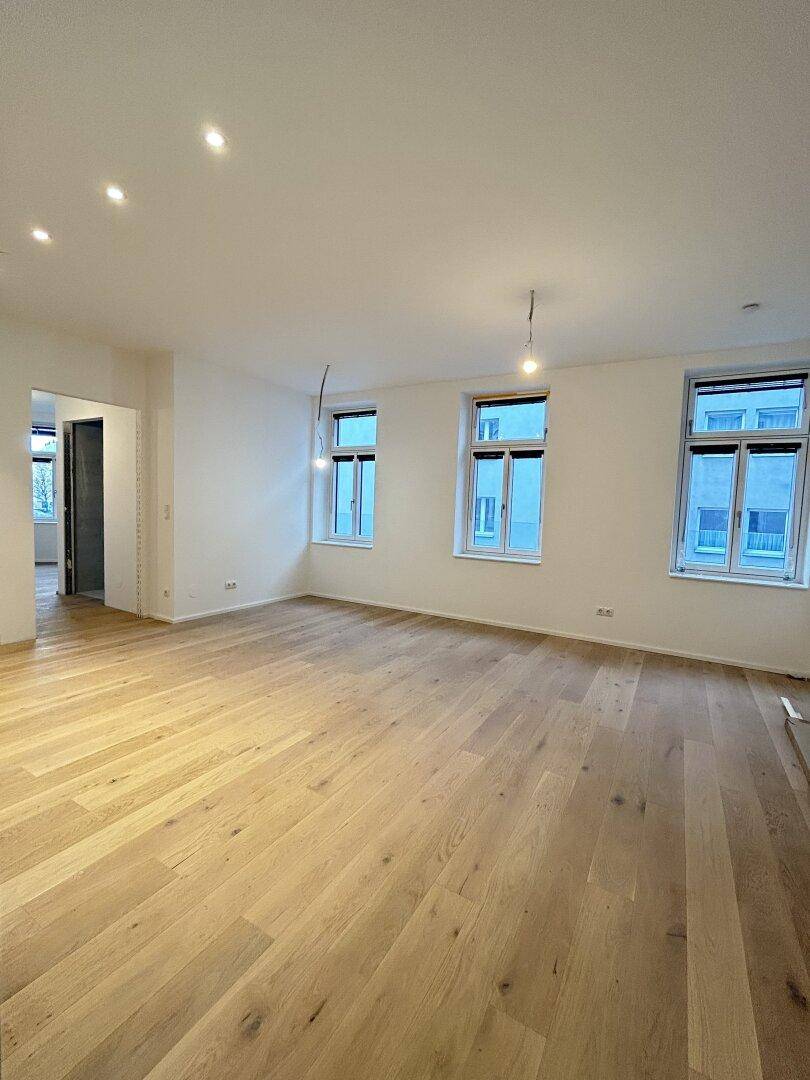 Geräumiger Wohnbereich mit hellem Holzparkett und drei großen Fenstern, die viel Licht hereinlassen.