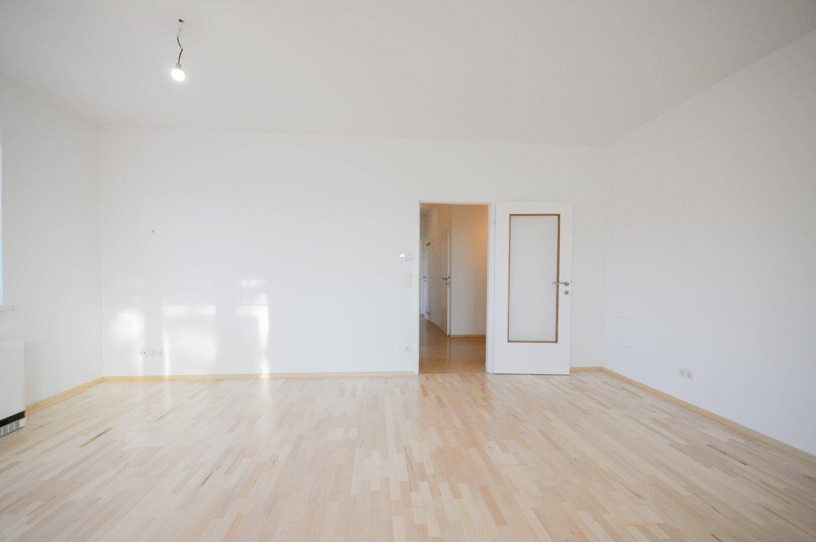 Geräumiges Wohnzimmer mit hellem Parkettboden und Zugang zu einem Flur, der weitere Räume verbindet.