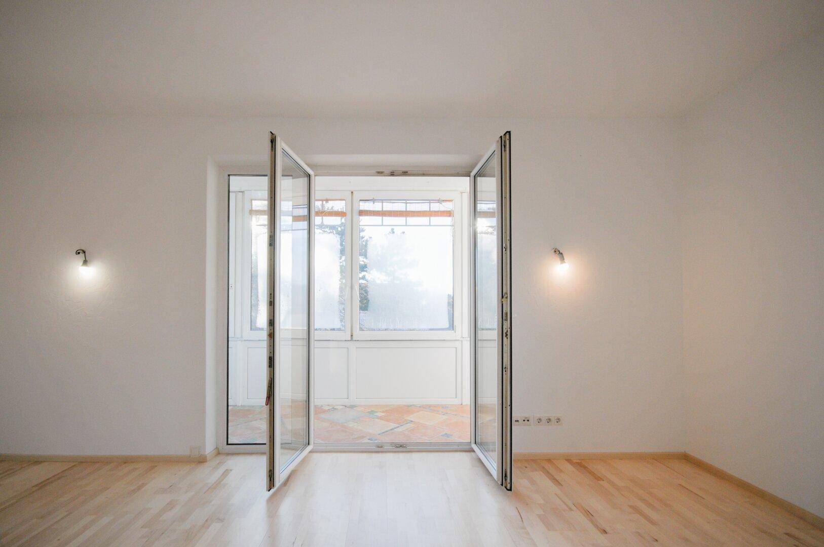 Heller Wohnraum mit Parkettboden und geöffneten Balkontüren, die viel Tageslicht hereinlassen.