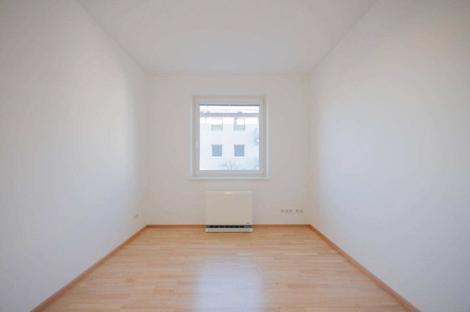 Helles Zimmer mit Parkettboden und Fenster, das einen Blick auf die umliegenden Gebäude bietet.