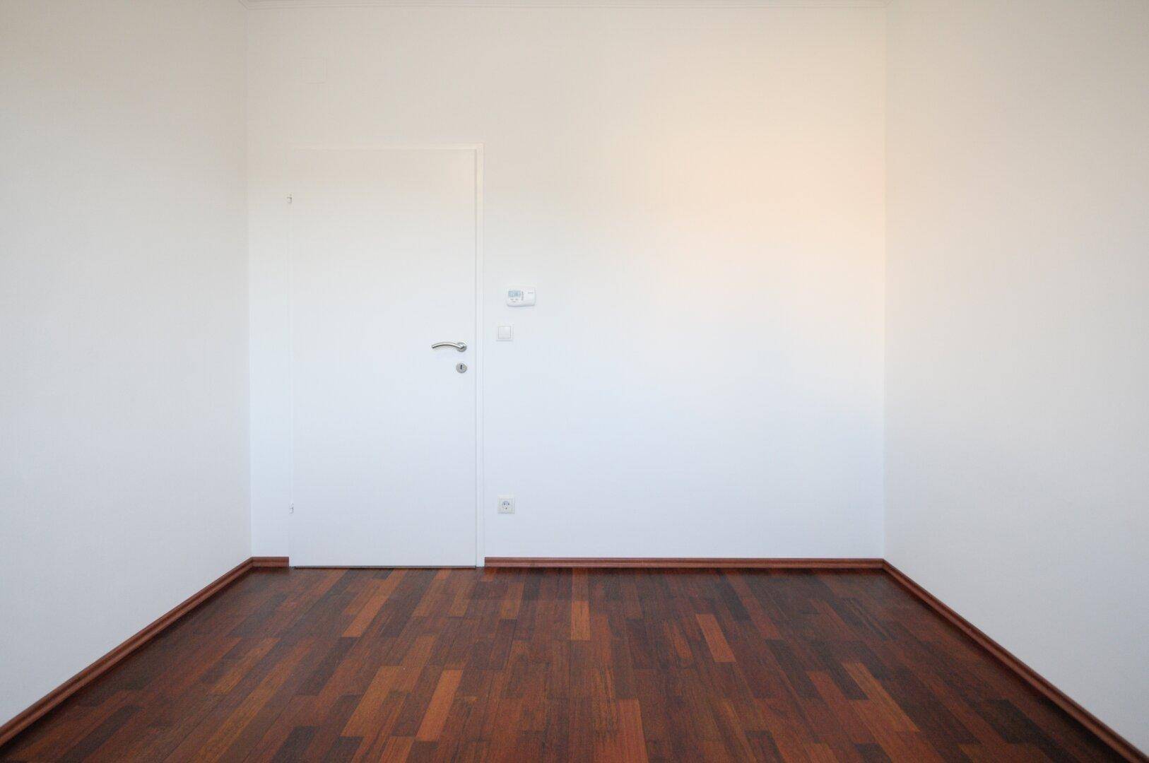 Leeres Zimmer mit dunklem Parkettboden und einer weißen Tür, bereit zur individuellen Einrichtung.