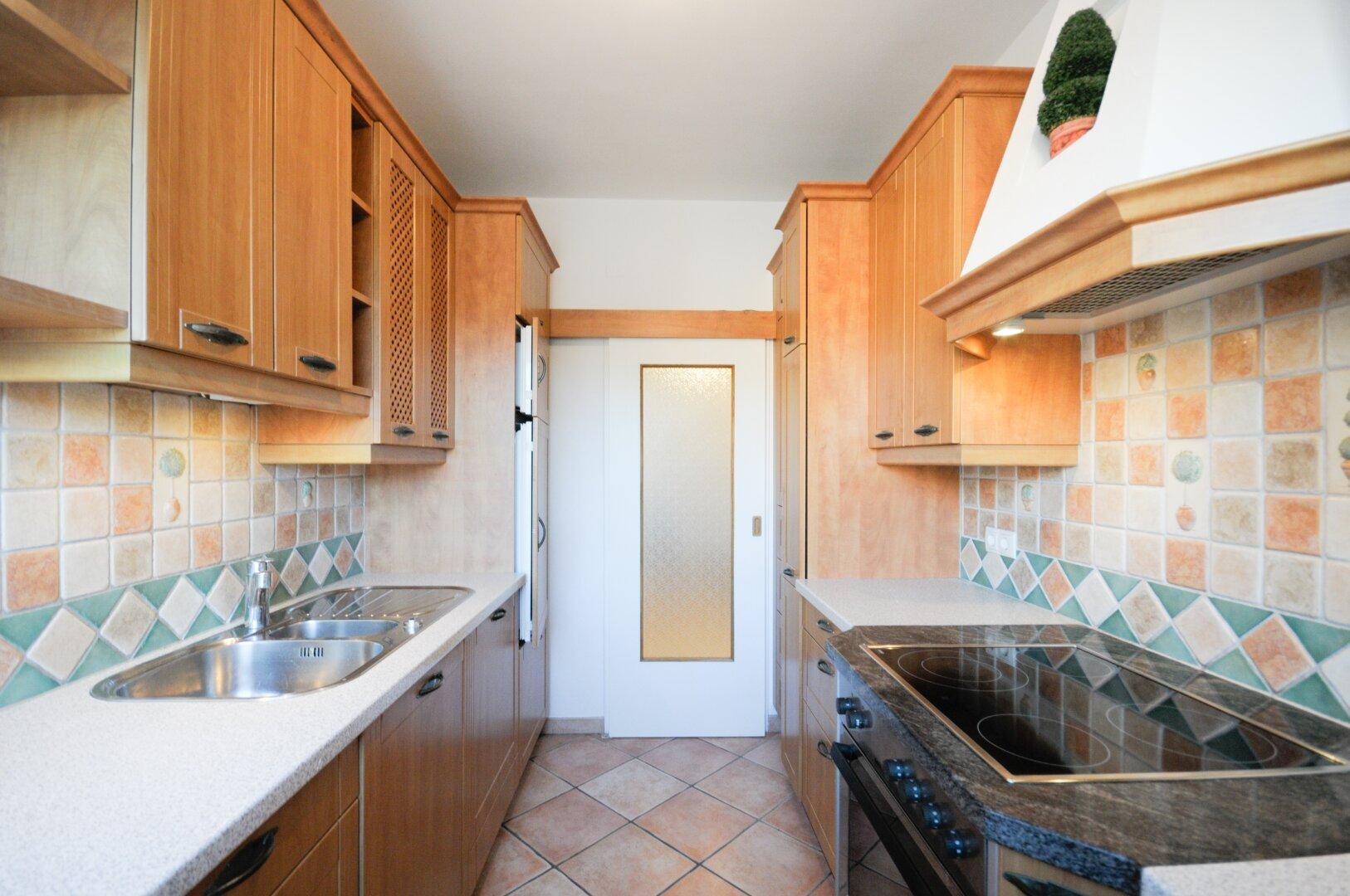 Funktionale Küche mit Holzschränken, Terrakotta-Fliesen und einer Tür, die zu weiteren Räumen führt.