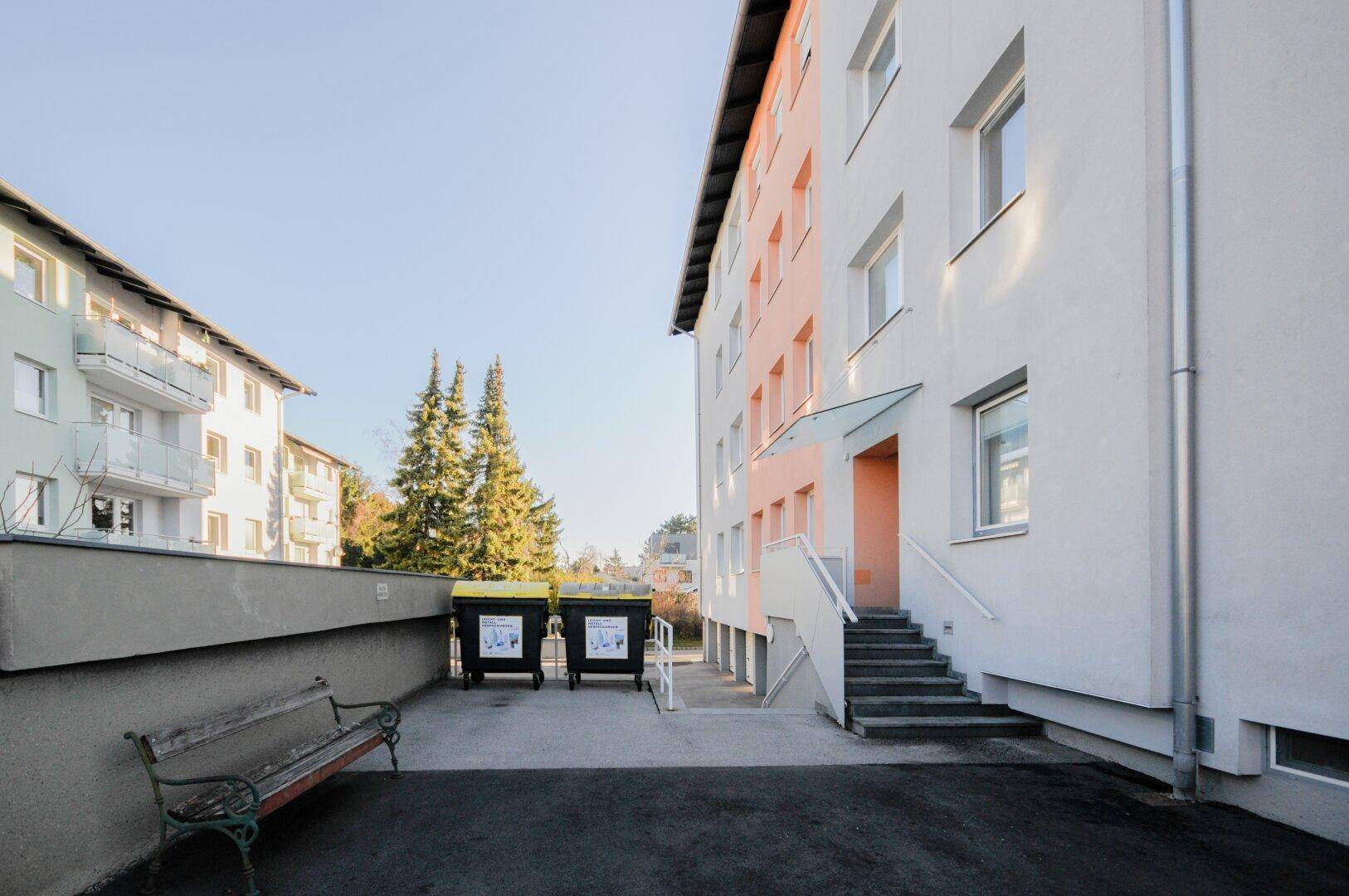 Außenansicht des Mehrfamilienhauses mit Mülltonnen und einer Bank im Vordergrund.