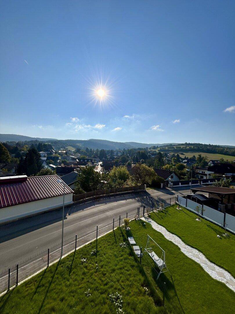 Sonniger Garten mit Rasen und gepflastertem Bereich, umgeben von ländlicher Landschaft.