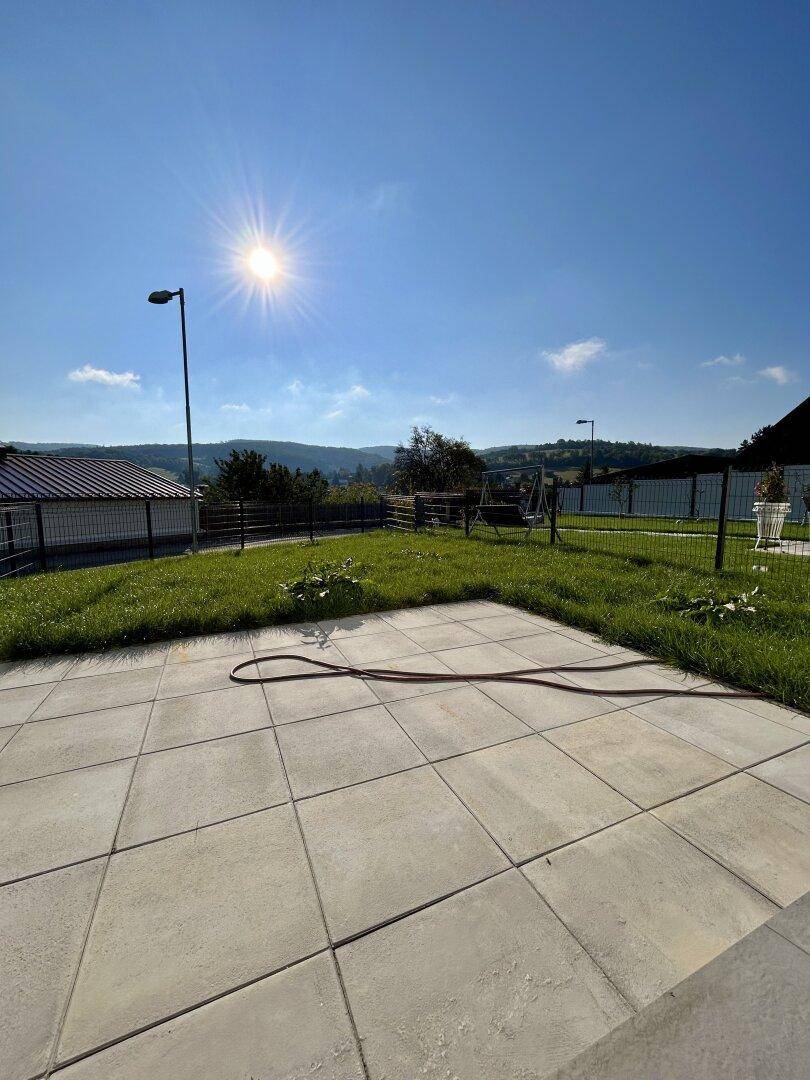 Sonniger Garten mit Rasenfläche und gepflasterter Terrasse, umgeben von Natur.