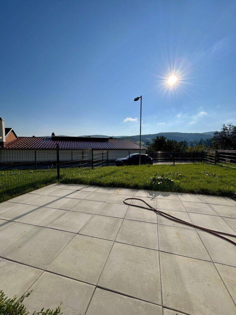 Sonniger Garten mit Rasenfläche und gepflasterter Terrasse, umgeben von Natur.